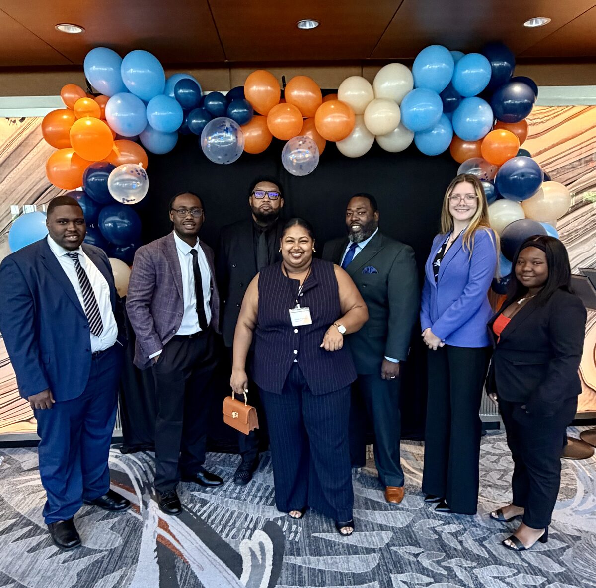 The ALL-IN Marketing team in business attire, standing together beneath an orange and blue balloon arch at a company event